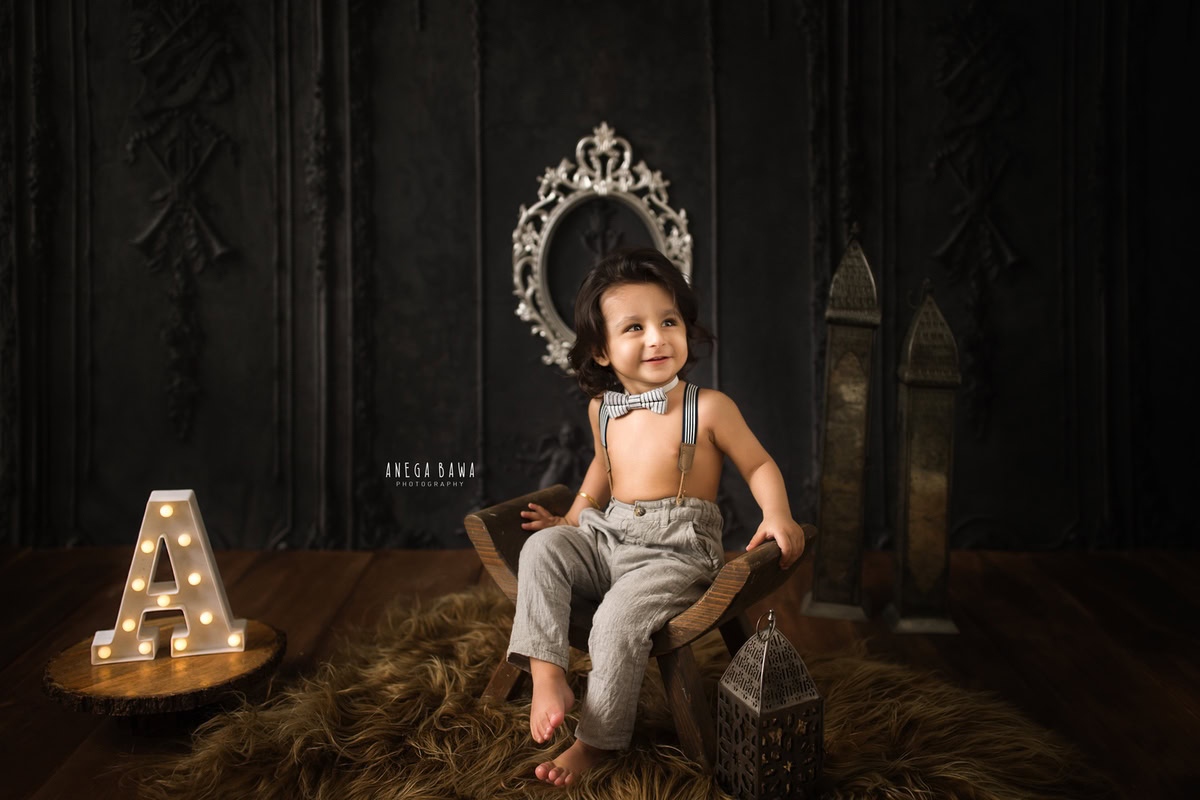 Capture the adorable moments of your 9-month to 1-year-old boy in a sitter photoshoot by Anega Bawa Photography in Delhi and Gurgaon. Against a warm brown backdrop with a silver frame on the wall, and seated on a slant wooden chair with a cute bow, these photos will be cherished memories of his early years.