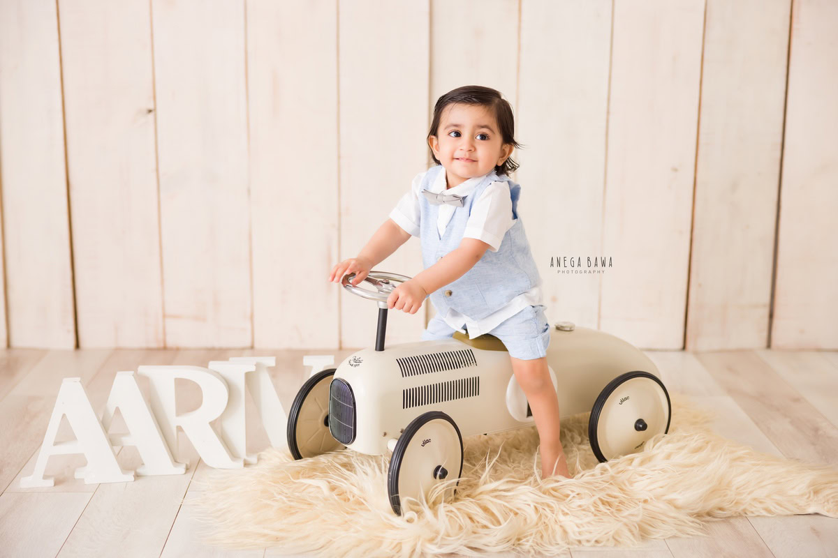 In Gurgaon, Anega Bawa, a talented sitter photographer, artfully documents a 9-month to 1-year-old boy seated on a toy scooter with a personalized name frame adorning the floor against a warm beige backdrop. This charming portrait captures the joy and playfulness of the child's early years, beautifully preserved by the skilled photographer.