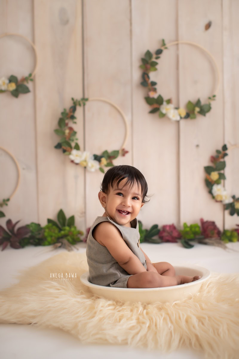 In Gurgaon, Anega Bawa, an adept sitter photographer, captures a serene moment of a 9-month to 1-year-old boy seated in a tub, surrounded by a cozy beige rug and set against a neutral beige backdrop. The scene is enhanced by a wooden leafy frame adorning the wall, adding a touch of natural elegance to the composition.