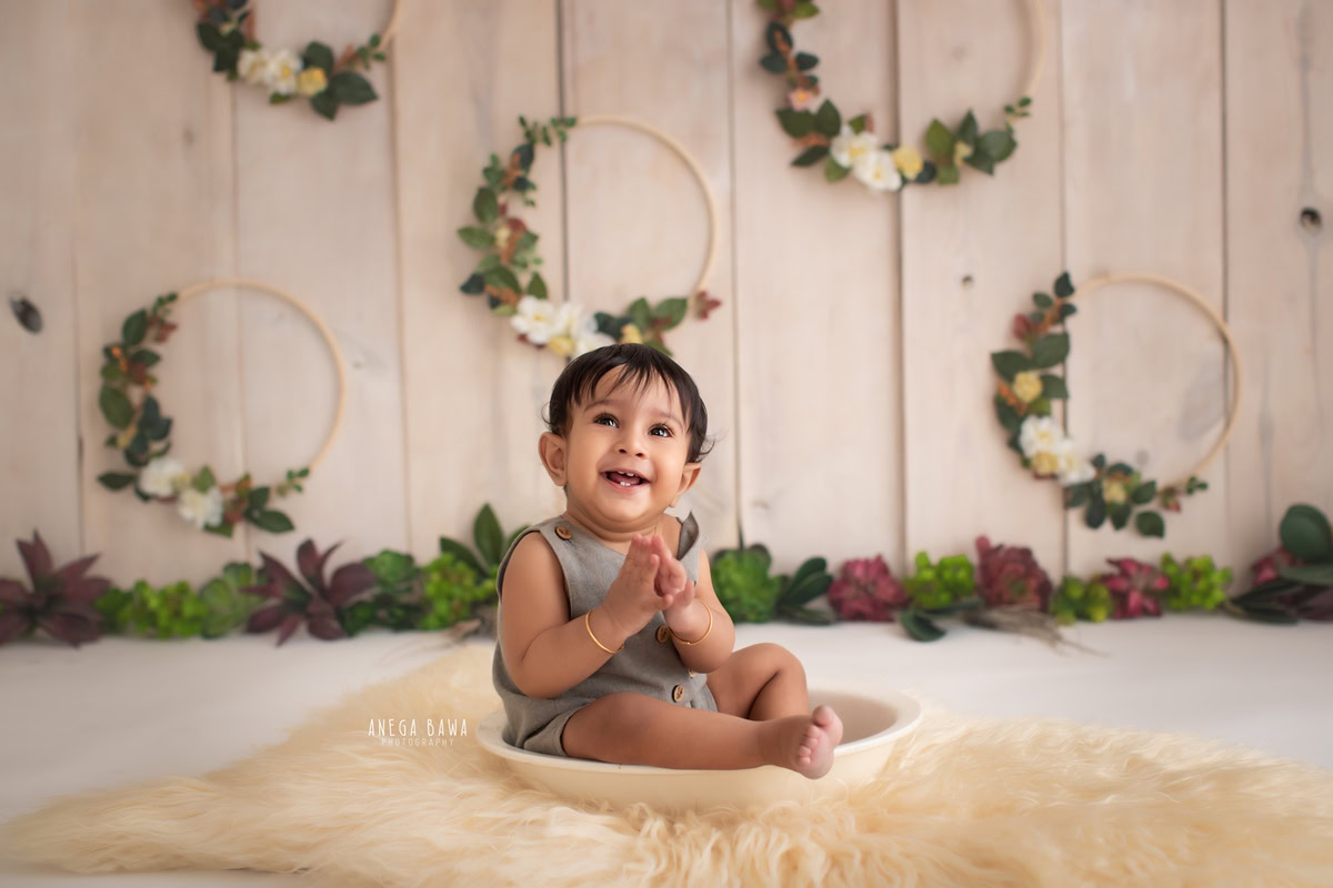 In Gurgaon, Anega Bawa, a skilled sitter photographer, frames a precious moment of a 9-month to 1-year-old boy seated in a tub, nestled on a soft beige rug. The scene is enriched by a wooden floral frame gracing the wall against a soothing beige backdrop, creating a charming and timeless composition.