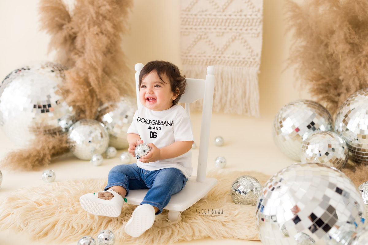 Capture the essence of your 9-month-old to 1-year-old boy with Anega Bawa Photography's sitter photography in Delhi and Gurgaon. Watch him seated on a white chair, surrounded by the whimsical allure of disco balls and bushes against a serene beige backdrop. Let every click encapsulate the innocence and wonder of his journey through infancy.