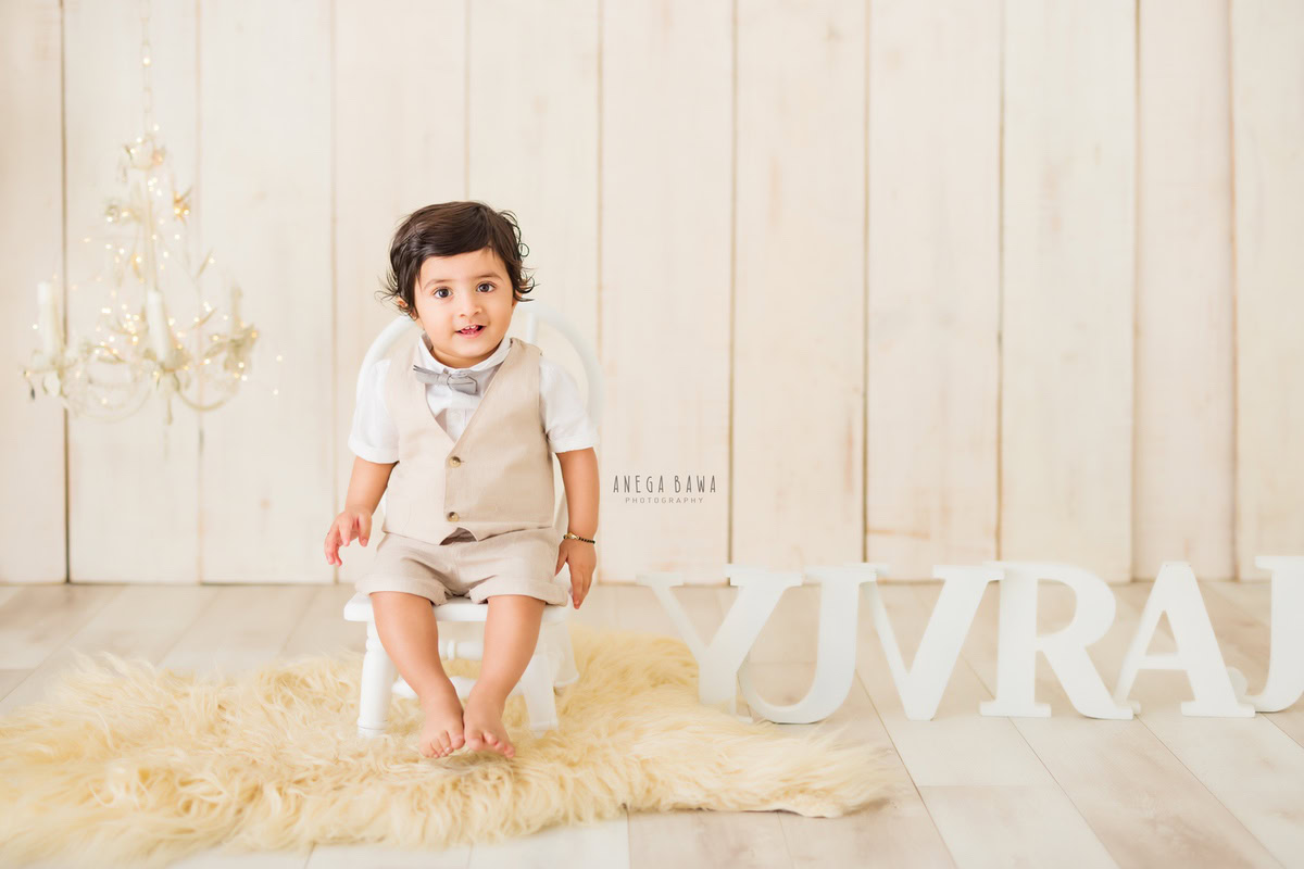 In this captivating image from a baby pre-birthday photoshoot by Anega Bawa in Delhi, a 9-month-old boy is seated on a white chair against a beige backdrop. The warm glow of candle stands and golden lights adds a magical touch to the scene, while a beige rug underneath provides comfort. A name frame on the floor adds a personalized element, contributing to the charm of the setup. It's a beautiful moment captured during this special milestone celebration.
