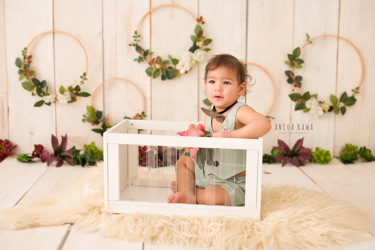 In this endearing sitter photography session by Anega Bawa in Delhi, Gurgaon, a 9-month-old boy is captured sitting on a white cot, nestled on a beige rug against a matching backdrop. The scene is adorned with floral wooden frames hung on the wall, adding a touch of rustic charm to the composition.