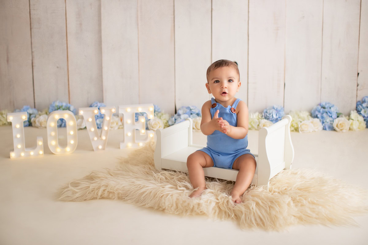 Create lasting memories of your 9-month to 1-year-old boy with a sitter photoshoot by Anega Bawa Photography in Delhi and Gurgaon. Seated on a white cot with a cozy beige rug beneath, surrounded by blue and white flowers and a love frame adorned with lights on the floor, these photos will capture the innocence and charm of his early years.
