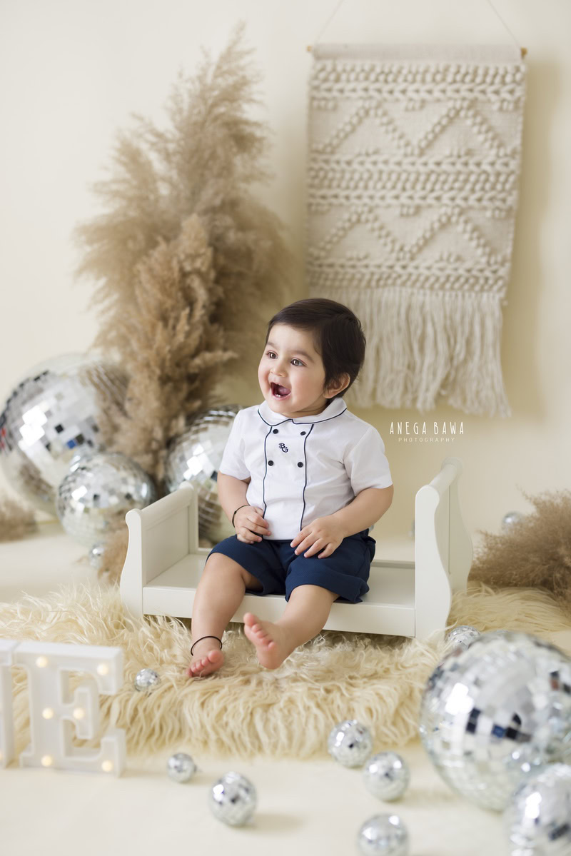In this enchanting capture by Gurgaon's talented sitter photographer, Anega Bawa, a boy aged 9 months to 1 year sits snugly on a white cot, surrounded by a beige rug. The scene is adorned with whimsical elements like discoballs and brown bushes, while fairy lights add a magical glow, creating a captivating ambiance.