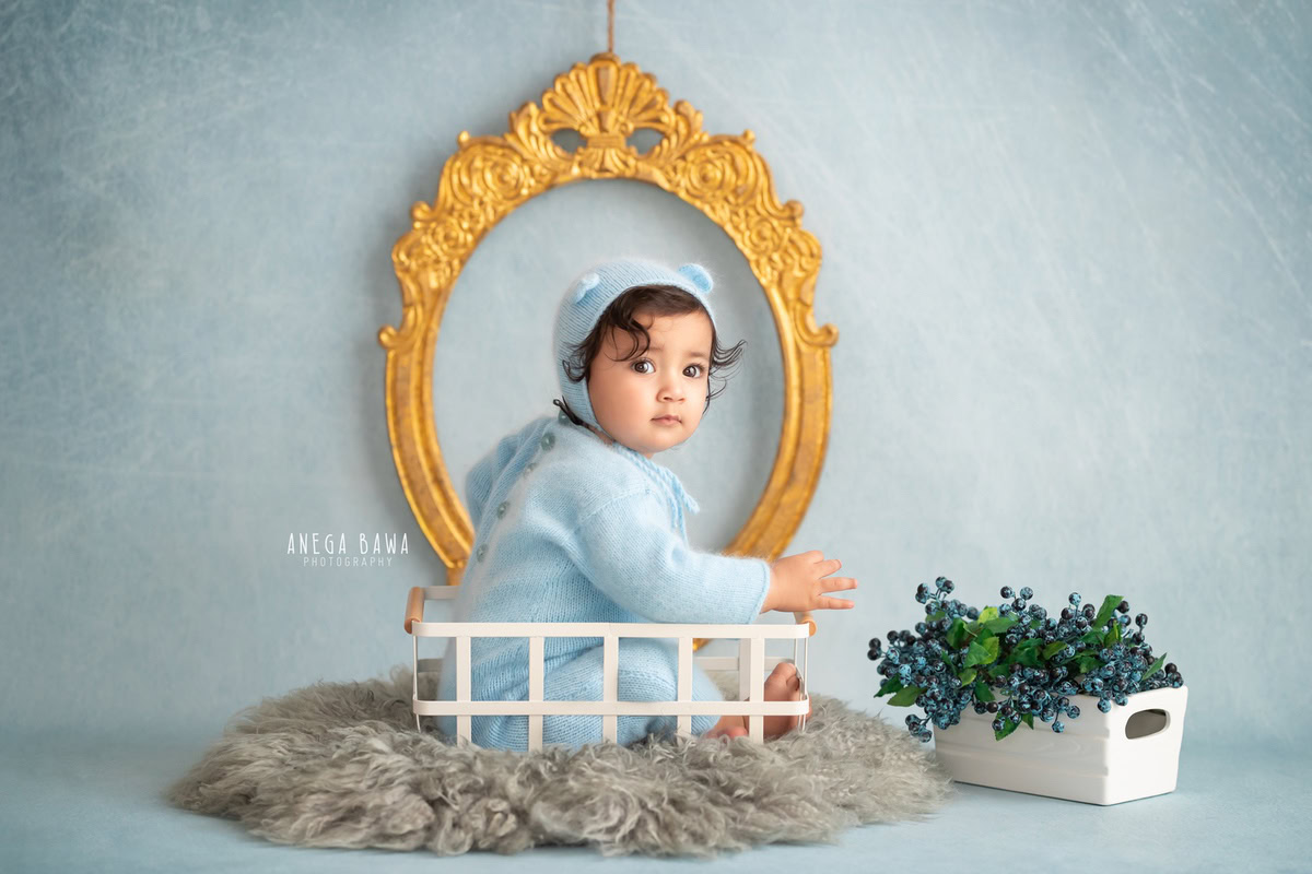 In this captivating photo captured by Gurgaon's skilled sitter photographer, Anega Bawa, a 9-month to 1-year-old boy sits serenely on a white cot against a striking blue backdrop. A golden frame on the wall adds a touch of elegance to the scene, creating a mesmerizing composition.