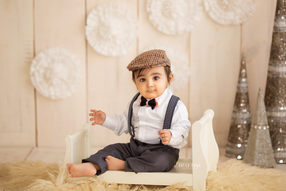Capture the precious moments of your 9-month to 1-year-old boy with a sitter photoshoot by Anega Bawa Photography in Delhi and Gurgaon. Seated on a white cot against a beige backdrop, adorned with a cute hat and a pompom on the wall, set on a cozy beige rug, these photos will cherish the innocence and joy of his early years.