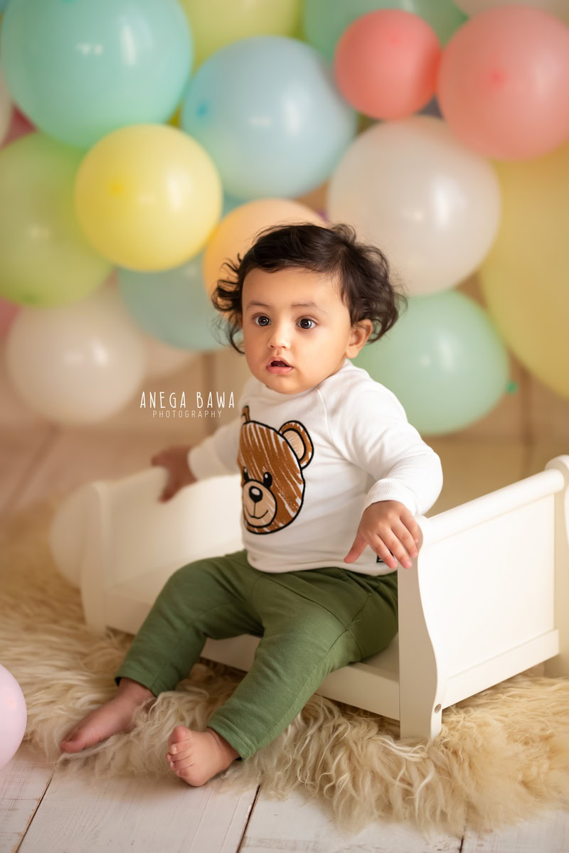 Create unforgettable memories of your 9-month to 1-year-old boy with a sitter photoshoot by Anega Bawa Photography in Delhi and Gurgaon. Picture him seated on a white cot surrounded by vibrant multicolored balloons, set on a cozy beige rug, capturing the joy and playfulness of his early years.