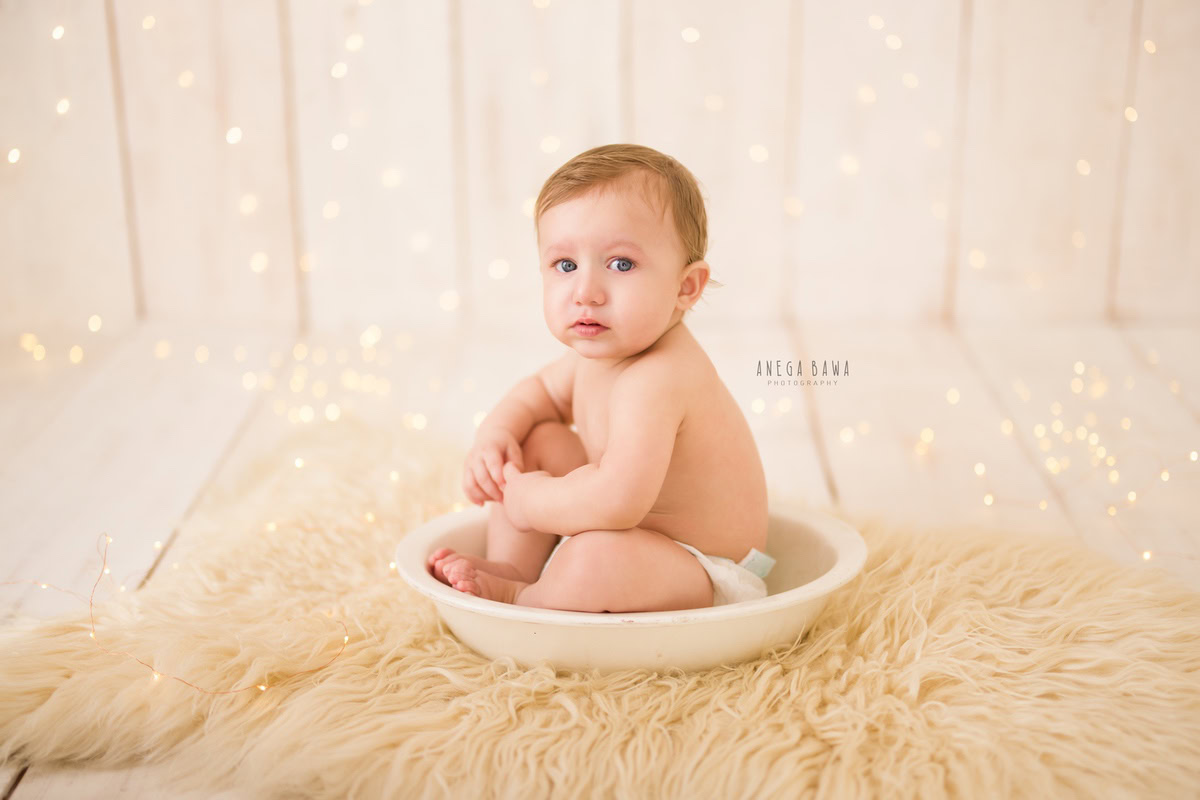 In this captivating sitter photography session by Anega Bawa in Delhi, Gurgaon, a 10-month-old boy is captured seated in a white tub, adorned with golden lights, placed on a cozy beige rug. Against the soothing beige backdrop, his innocence shines through, creating a timeless moment to cherish.
