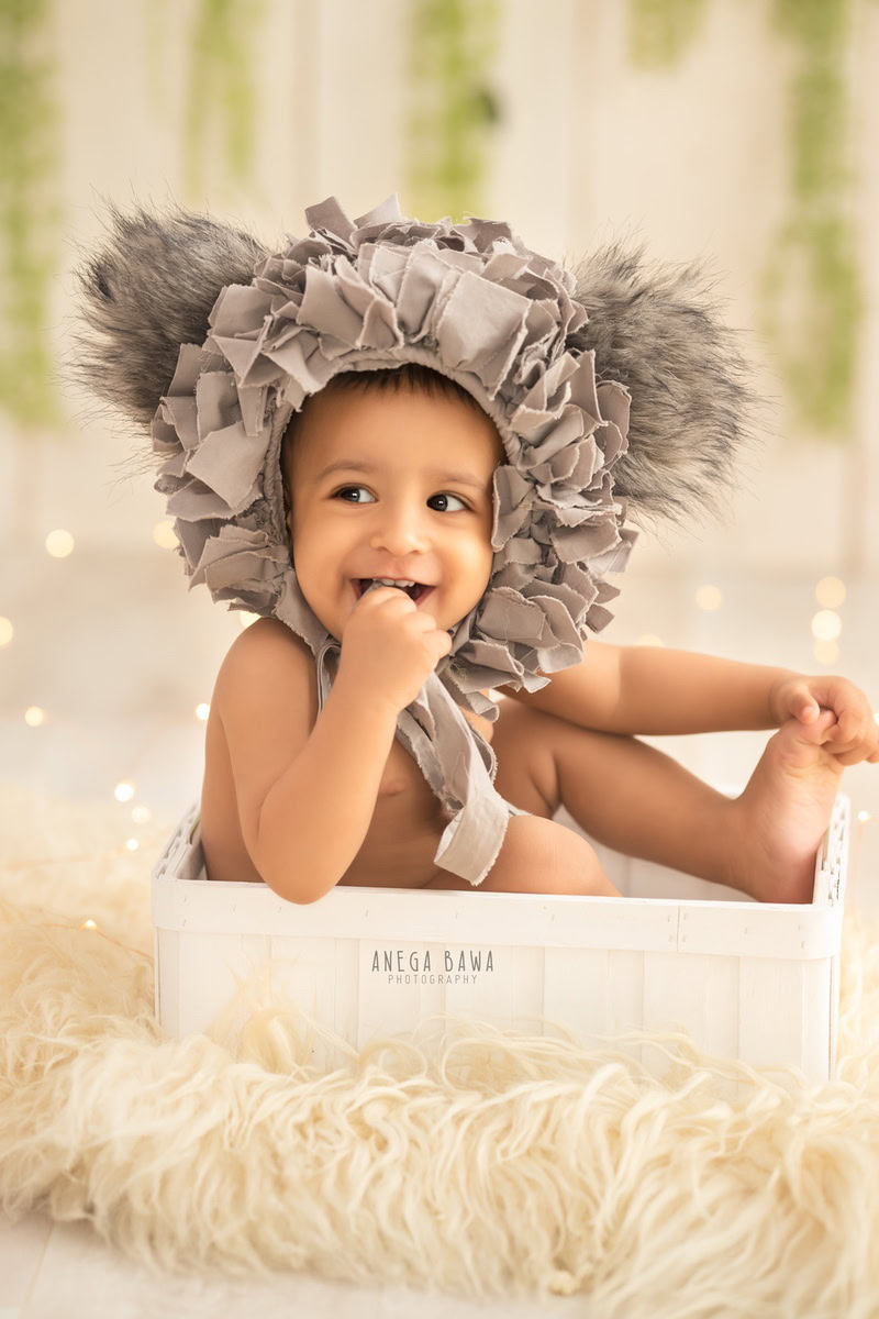 In this enchanting capture by Gurgaon's talented sitter photographer, Anega Bawa, a 10-month to 1-year-old boy sits in a white tub adorned with a unique cap, adding a whimsical touch to the scene. Against a beige backdrop, the boy is surrounded by a cozy atmosphere accentuated by a beige rug and leafy fringes on the wall, creating a delightful composition.