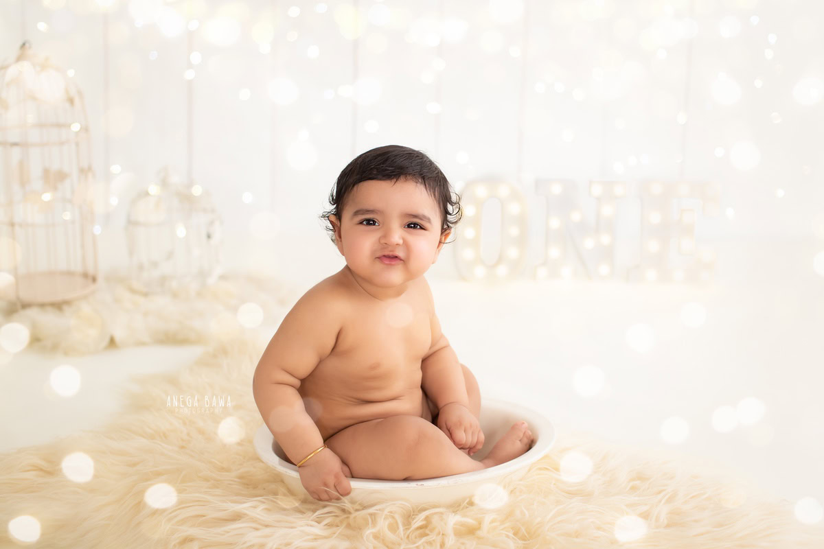 Captured by Anega Bawa, a prominent sitter photographer in Gurgaon, this enchanting image features a 10-month to 1-year-old boy seated in a white tub. Delicate fairy lights illuminate the scene, casting a soft glow as the boy interacts with charming white castles. The minimalist beige backdrop, expertly curated by Anega Bawa, adds a touch of sophistication to the composition.