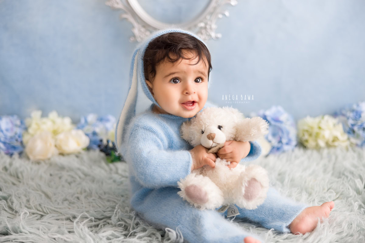 Captured by renowned sitter photographer Anega Bawa in Delhi, this delightful image showcases a 10-month to 1-year-old boy seated on a grey rug, adorned in a charming blue baby suit. He is accompanied by a cuddly soft toy as he interacts with beautiful blue and white flowers. The expertly curated composition exudes warmth and innocence, making it a perfect keepsake of this precious moment.