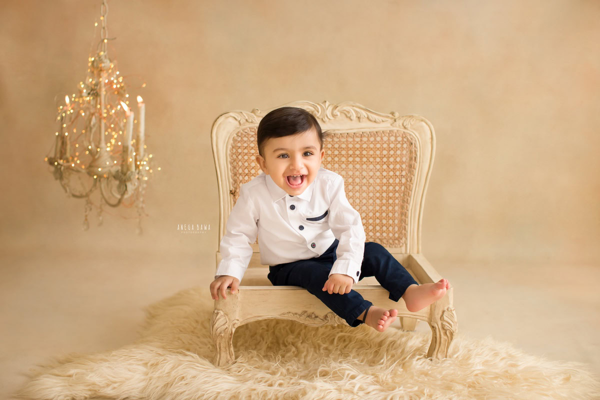 Create timeless memories of your 10-month to 1-year-old boy with a sitter photoshoot by Anega Bawa Photography in Delhi and Gurgaon. Let him sit on a wooden chair, surrounded by a cozy beige rug, with candle stands and golden lights adding warmth against a matching beige backdrop, ensuring an enchanting ambiance for the photoshoot.