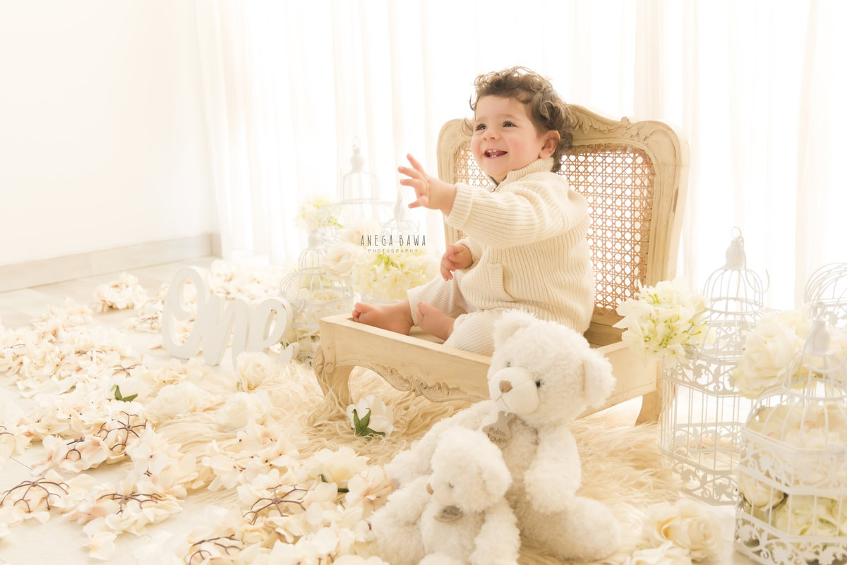 Boy seated on a wooden chair amidst white castles, flowers on the floor, soft toy, white backdrop - 10 months to 1-year-old first birthday photoshoot in Delhi by Anega Bawa, Gurgaon, Noida
