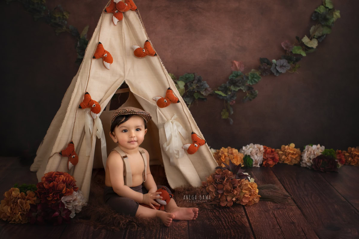 In this captivating photo captured by the talented sitter photographer Anega Bawa in Gurgaon, a 10-month to 1-year-old boy is seated on a wooden floor, surrounded by an adorable tent adorned with orange and white flowers. The rustic brown backdrop adds depth to the scene, creating a charming and whimsical atmosphere. This expertly composed image beautifully captures the innocence and wonder of childhood, making it a cherished memory for years to come.