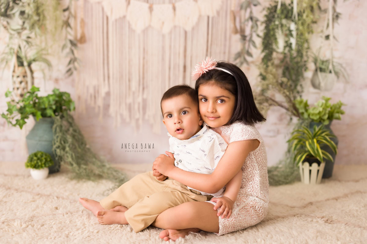 Boy with his sibling, seated on a plush beige rug beside a verdant green pot adorned with lush leaves, against a serene beige backdrop - capturing the warmth and joy of their 11 months to 1-year-old first birthday photoshoot in Delhi by Anega Bawa, Gurgaon, Noida.