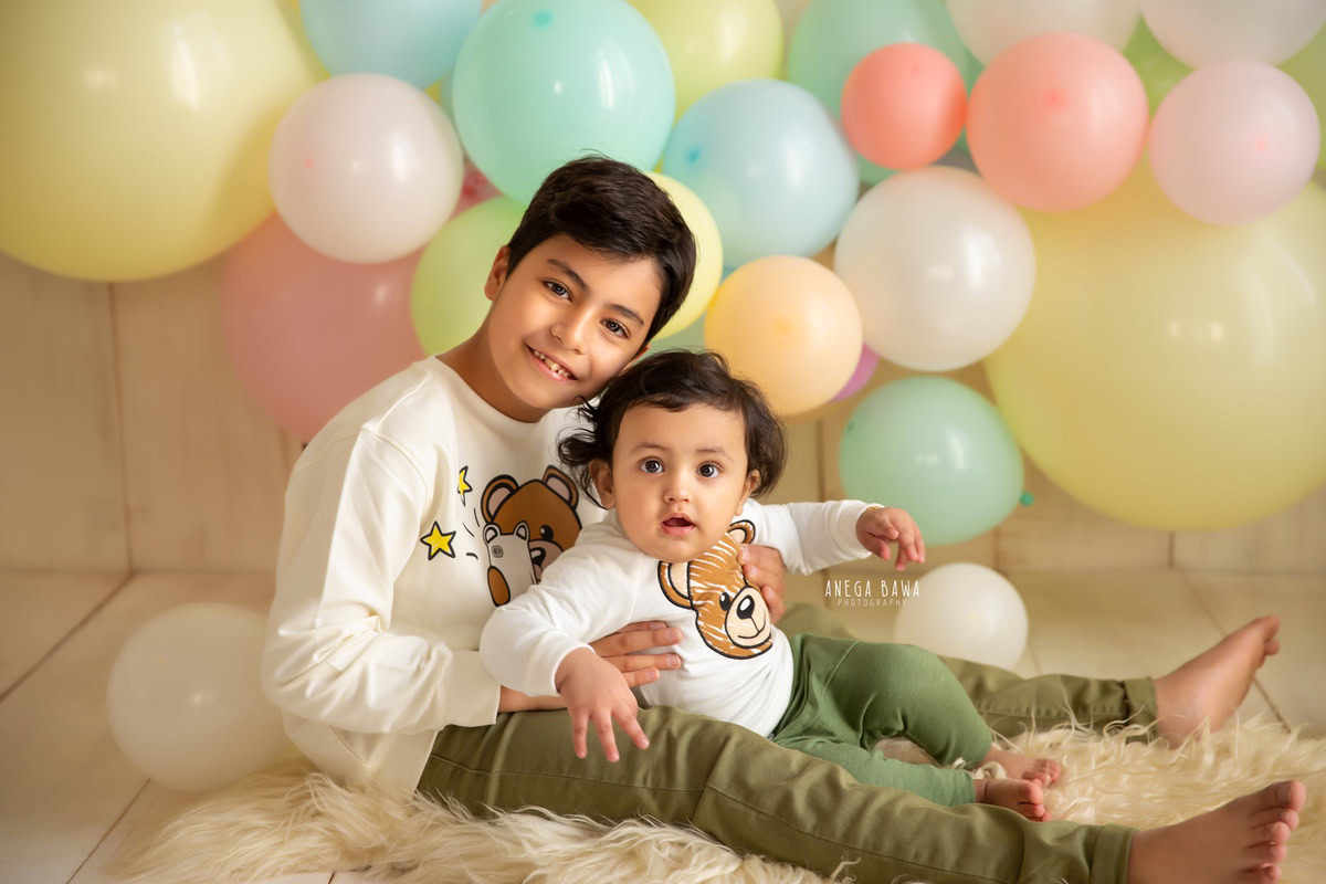 Capture heartwarming moments of your 11-month to 1-year-old boy and his sibling with a sitter photoshoot by Anega Bawa Photography in Delhi and Gurgaon. Set against a neutral beige backdrop, let them sit on a comfy beige rug surrounded by colorful balloons, creating joyous memories to cherish forever.