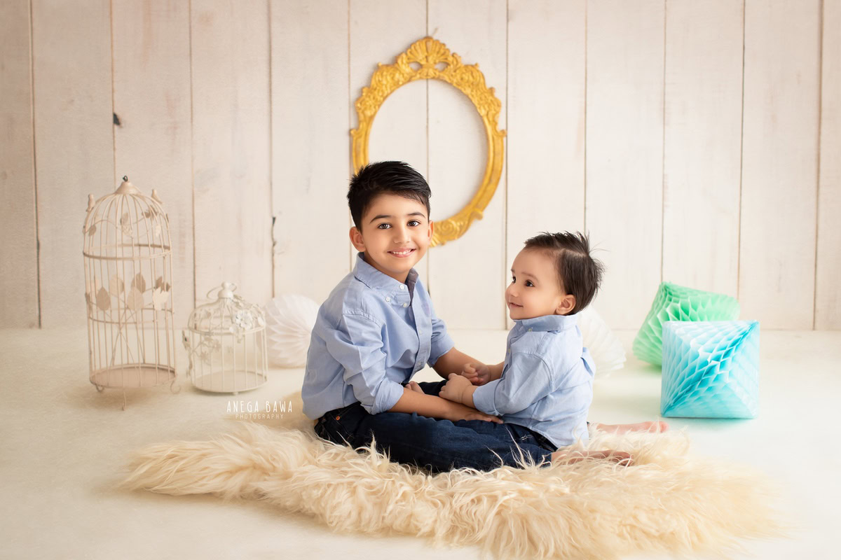 Boy with his sibling, framed by a golden frame on the wall against a serene beige backdrop, surrounded by colorful pompoms, marking their transition from 11 months to 1 year old during their first birthday photoshoot in Delhi by Anega Bawa, Gurgaon, Noida.