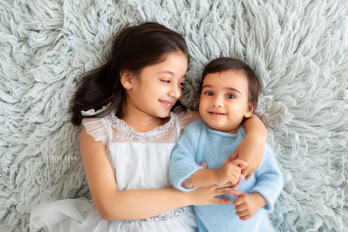 In this endearing photograph captured by the skilled sitter photographer Anega Bawa in Gurgaon, two siblings aged 11 months to 1 year old share an adorable hugging pose on a soft grey rug. Their sweet embrace radiates warmth and affection, creating a heartwarming moment frozen in time.