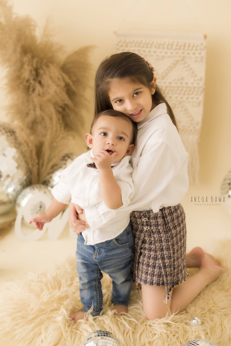 Boy and his sibling sharing a heartwarming hug on a soft beige rug amidst lush brown bushes and a sparkling disco ball, against a tranquil beige backdrop, capturing the tender bond between them during their 11 months to 1-year-old first birthday photoshoot in Delhi by Anega Bawa, Gurgaon, Noida.