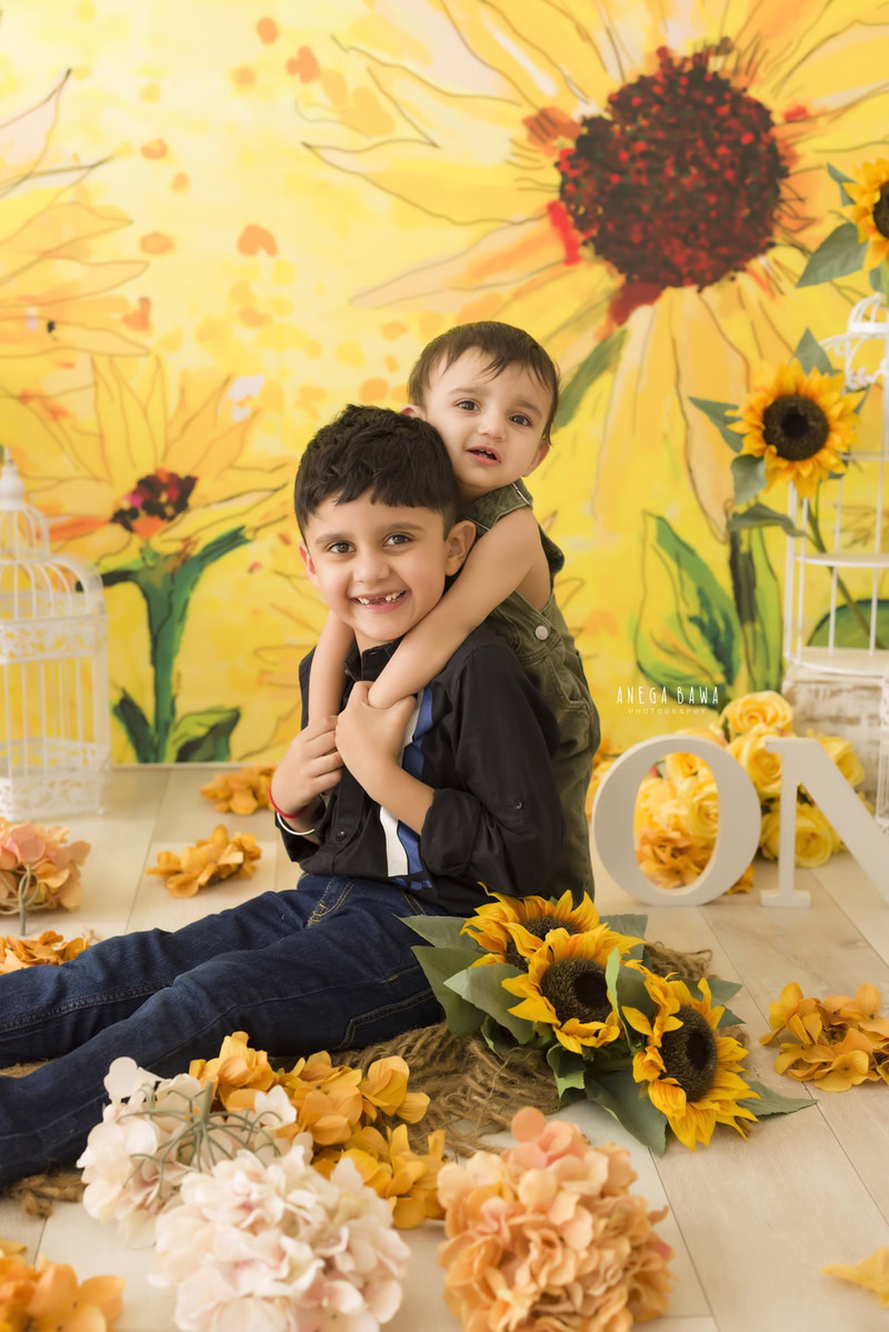 Boy with his sibling in front of a radiant sunflower backdrop surrounded by vibrant orange flowers, commemorating their journey from 11 months to 1 year old during their first birthday photoshoot in Delhi by Anega Bawa, Gurgaon, Noida.