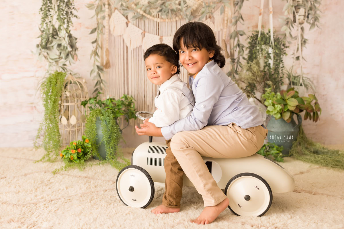 In this captivating image taken by the talented sitter photographer Anega Bawa in Delhi, two siblings aged 11 months to 1 year old are seen with a toy scooter against a charming beige net backdrop. The scene is adorned with delightful castles and lush green leafy fringes, adding a touch of whimsy and wonder to the photo.