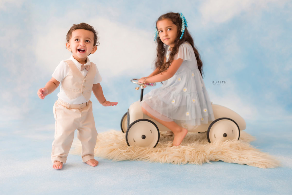 Amidst a whimsical backdrop reminiscent of fluffy clouds, two siblings, aged 11 months and 1 year old, enjoy a playful moment with a toy scooter. This heartwarming scene, captured during a baby pre-birthday photoshoot in Delhi by the talented Anega Bawa, showcases the innocence and joy of childhood.