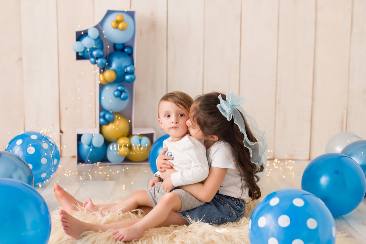 Adore the heartwarming bond between two siblings as they share a sweet kiss amidst a backdrop of blue balloons and a beige rug. These precious moments were captured during a toddler photoshoot in Delhi, Gurgaon, and Noida by the talented photographer, Anega Bawa, when the children were 11 months to 1 year old.