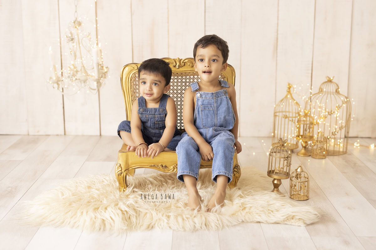 Captivating siblings dressed in denim dungarees, seated on a regal golden chair amidst golden castles on a cozy beige rug against a serene beige backdrop, marking their transition from 11 months to 1 year old during their first birthday photoshoot in Delhi by Anega Bawa, Gurgaon, Noida.