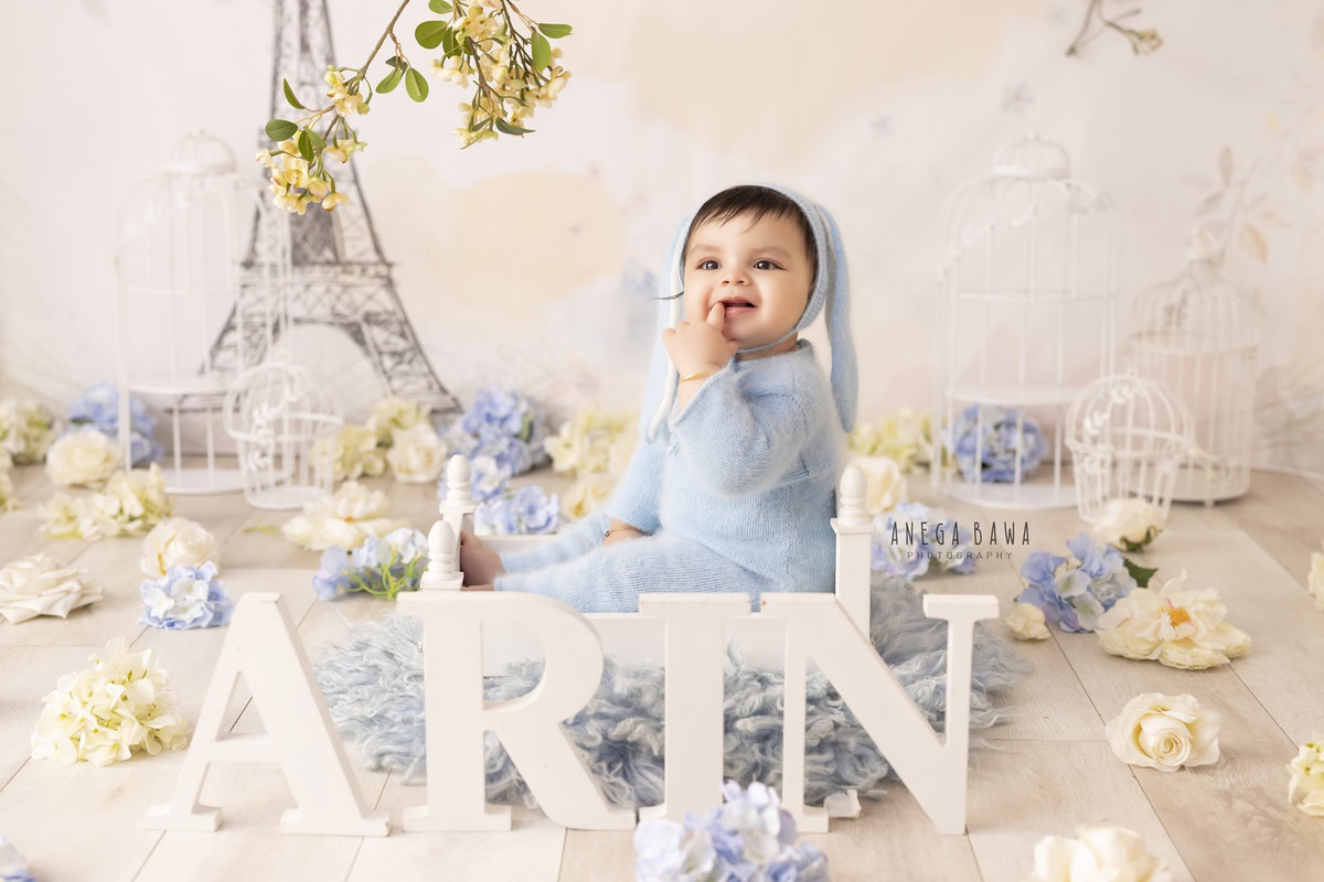In this adorable toddler photoshoot by Anega Bawa in Delhi, Gurgaon, and Noida, a charming little boy sits comfortably in a blue babysuit and a cute cap. The backdrop features a picturesque Eiffel Tower wallpaper, complemented by flowers scattered on the floor and a cozy blue rug. These captivating moments were captured when the boy was 11 months to 1 year old.