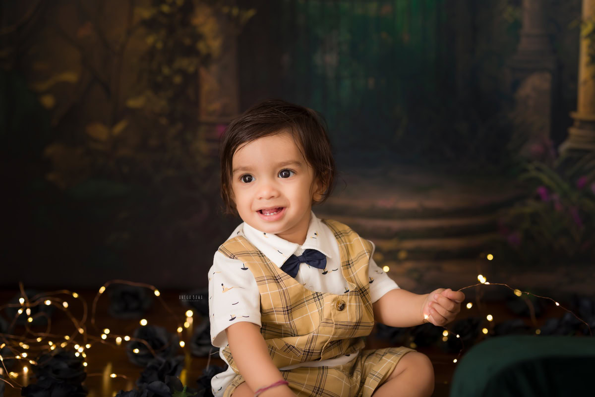 Boy sitting against a warm brown backdrop, illuminated by golden lights, commemorating his journey from 12 months to 1 year old during his first birthday photoshoot in Delhi by Anega Bawa, Gurgaon, Noida.