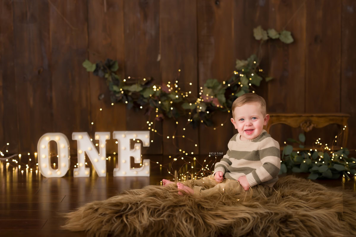 Amidst a cozy setting of earthy tones, with a rich brown rug against a matching backdrop, delicate fairy lights illuminate the scene, casting a magical glow around the adorable 12-month-old boy. Captured by the talented photographer Anega Bawa, this captivating moment during his baby pre-birthday photoshoot in Delhi radiates warmth and charm, preserving the joyous essence of childhood.