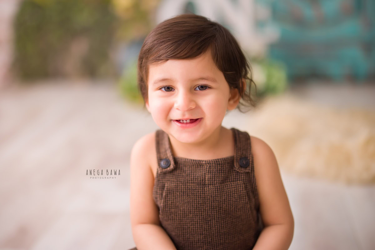Captured skillfully by the expert sitter photographer Anega Bawa in Gurgaon, this image features an adorable 12-month-old boy seated on a cozy beige rug, dressed in charming brown dungarees.