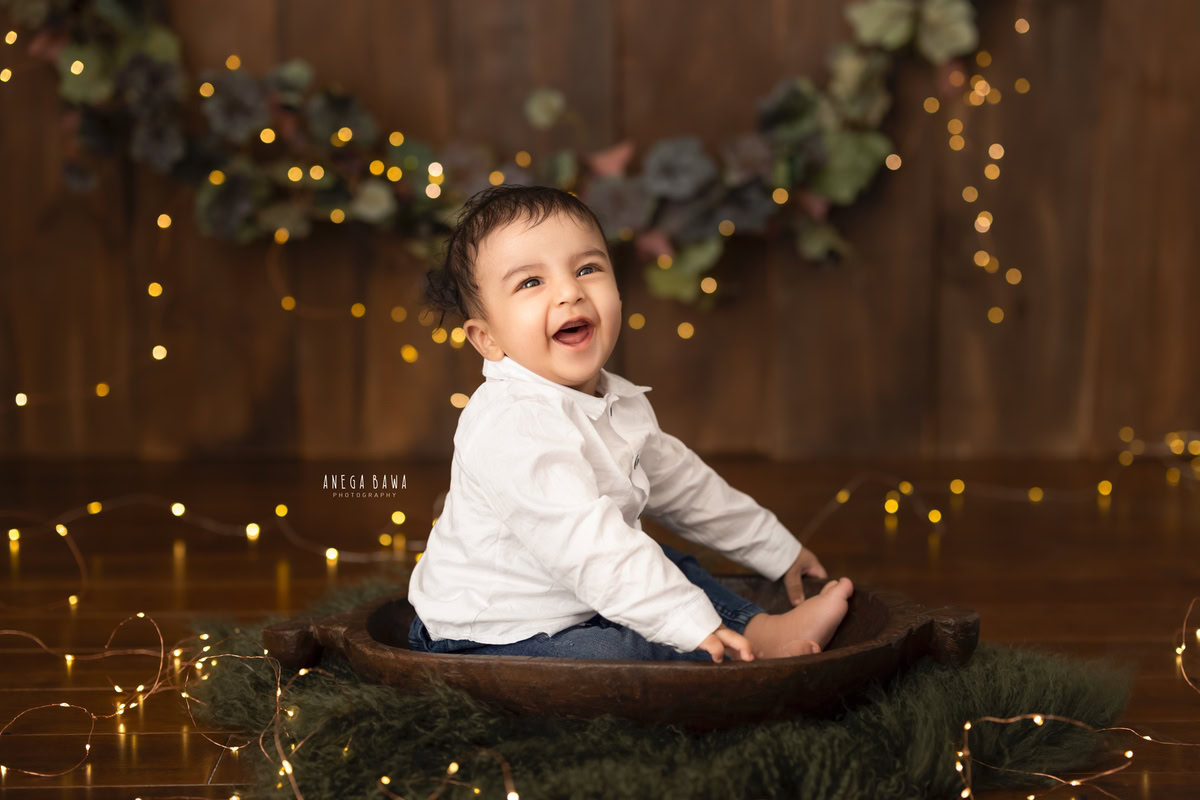 In this captivating sitter photography session in Delhi, Gurgaon by Anega Bawa, a 12-month-old boy sits comfortably in a brown tub surrounded by enchanting fairy lights. The scene is accentuated by the delicate leaves fringe adorning the wall, creating a whimsical ambiance. This magical setup perfectly captures the innocence and wonder of childhood, preserving precious memories for years to come.