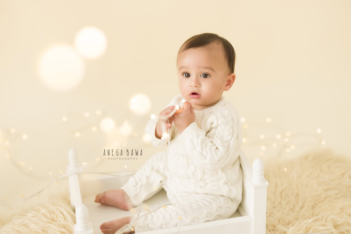 In this delightful toddler photoshoot by Anega Bawa in Delhi, Gurgaon, and Noida, a charming little boy, aged 12 months (1 year old), sits cozily in a white cot adorned with a beige rug. The serene beige backdrop is illuminated by twinkling fairy lights, adding a magical touch to the scene.