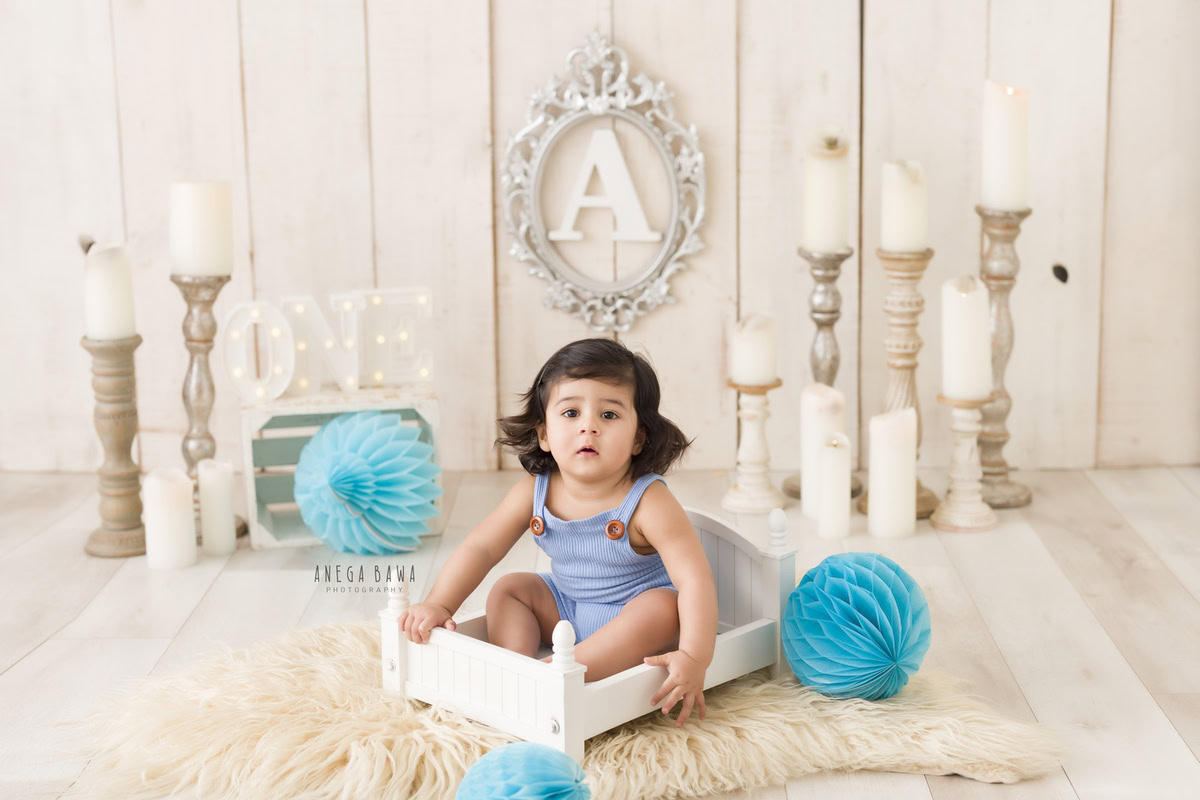 In this adorable toddler photoshoot captured by Anega Bawa in Delhi, Gurgaon, and Noida, a sweet little boy, aged 12 months (1 year old), sits comfortably in a white cot adorned with a beige rug. The scene is beautifully complemented by a candle stand and an alphabet frame on the wall. Delicate blue paper flowers and pompoms add a whimsical touch to the backdrop, creating a charming atmosphere.