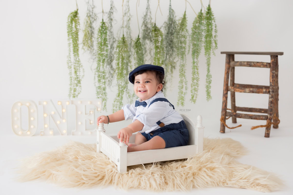 Boy seated in a pristine white cot on a cozy beige rug, with a leafy fringe on the wall against a pristine white backdrop, capturing the essence of his journey from 12 months to 1 year old during his first birthday photoshoot in Delhi by Anega Bawa, Gurgaon, Noida.