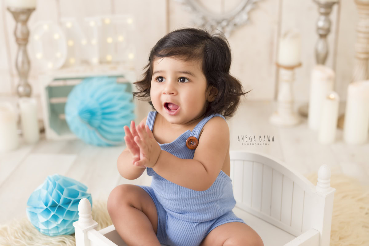 In this delightful toddler photoshoot by Anega Bawa in Delhi, Gurgaon, and Noida, a charming 12-month-old boy sits contentedly in a white cot. The scene is adorned with a quaint candle stand and embellished with blue paper flowers and pompoms. The little one looks adorable in a blue babysuit, adding to the enchanting ambiance of the photo.