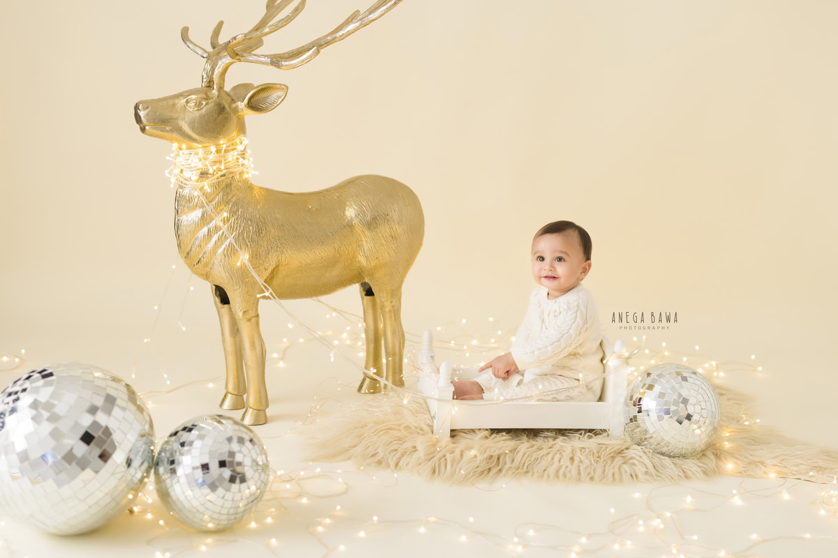 In this captivating toddler photoshoot by Anega Bawa in Delhi, Gurgaon, and Noida, a 12-month-old boy sits serenely in a white cot. The backdrop features a mesmerizing arrangement of golden deer and disco balls against a beige backdrop, creating a magical ambiance. The scene is further illuminated by fairy lights, adding a touch of enchantment to the photo.
