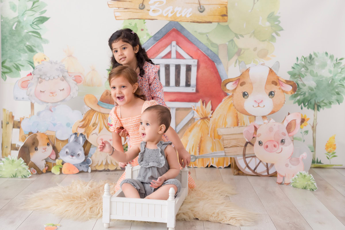 Boy seated in a pristine white cot with his siblings against a whimsical cartoon backdrop, commemorating his journey from 12 months to 1 year old during his first birthday photoshoot in Delhi by Anega Bawa, Gurgaon, Noida.
