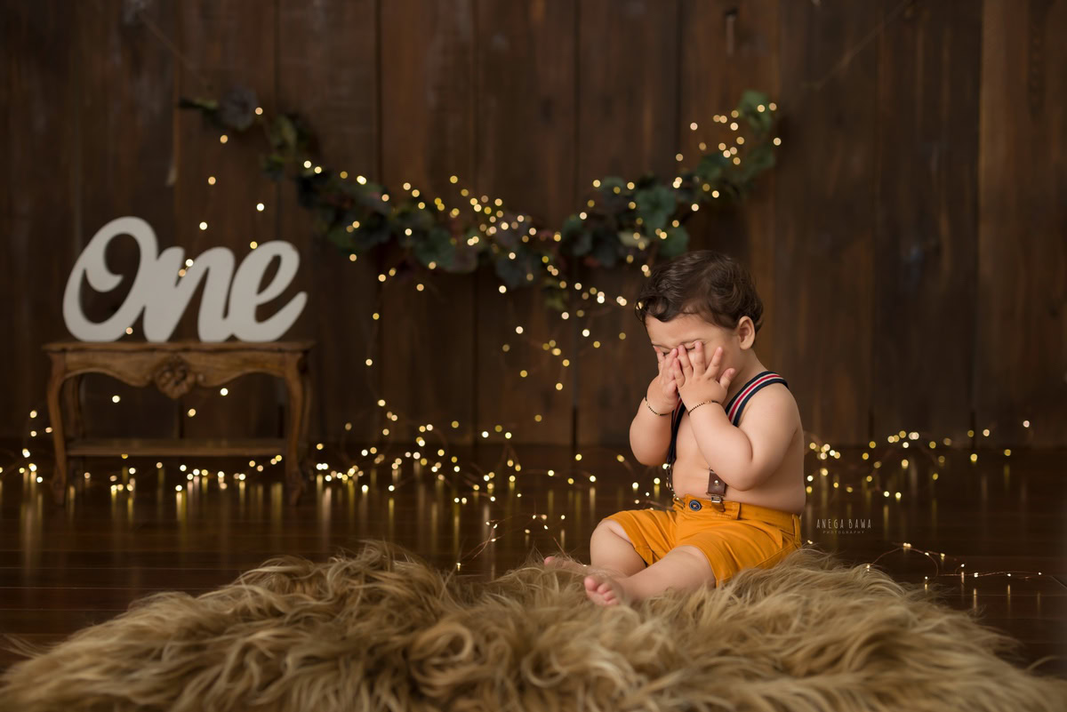 Capture the precious moments of your little boy's journey into toddlerhood with Anega Bawa Photography's sitter photography in Delhi and Gurgaon. Watch him sitting comfortably on a beige rug, surrounded by the warm glow of fairy lights against a serene brown backdrop. These timeless images will serve as cherished mementos of his playful spirit and growing personality at 12 months, making every click a treasure to behold.