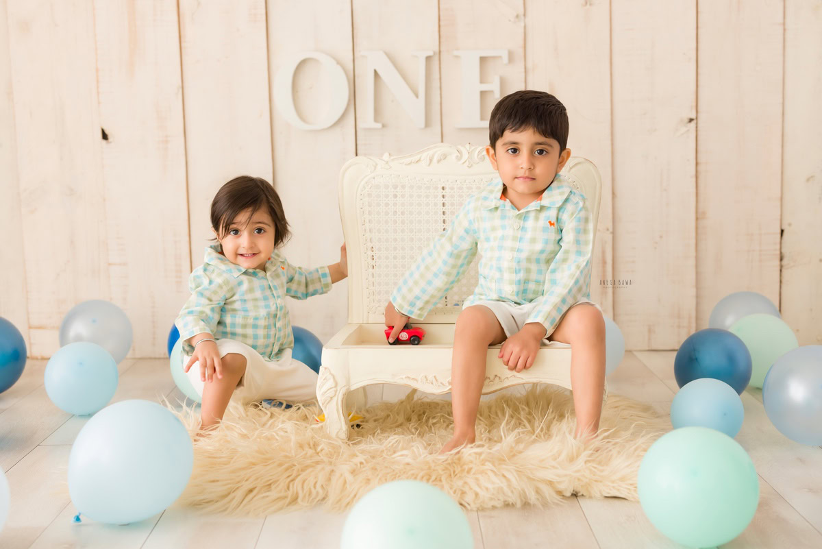 In this delightful sitter birthday photoshoot by Anega Bawa in Delhi, a 12-month-old boy is joined by his sibling as they sit on a beige rug against a serene beige backdrop adorned with balloons. The white chair provides a charming contrast to the beige elements, adding a touch of elegance to the scene. The siblings' joyful expressions and the colorful balloons create a cheerful atmosphere, capturing the essence of celebration and togetherness on this special occasion.