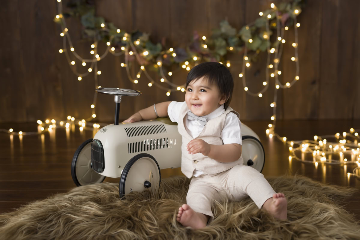 Witness the enchantment unfold as your 12-month-old boy explores the world around him in a sitter photoshoot by Anega Bawa Photography in Delhi and Gurgaon. Nestled on a warm brown rug against a rustic brown backdrop adorned with golden lights, he exudes curiosity and charm. With a toy scooter by his side, each snapshot captures his playful spirit and budding personality, creating cherished memories to last a lifetime.