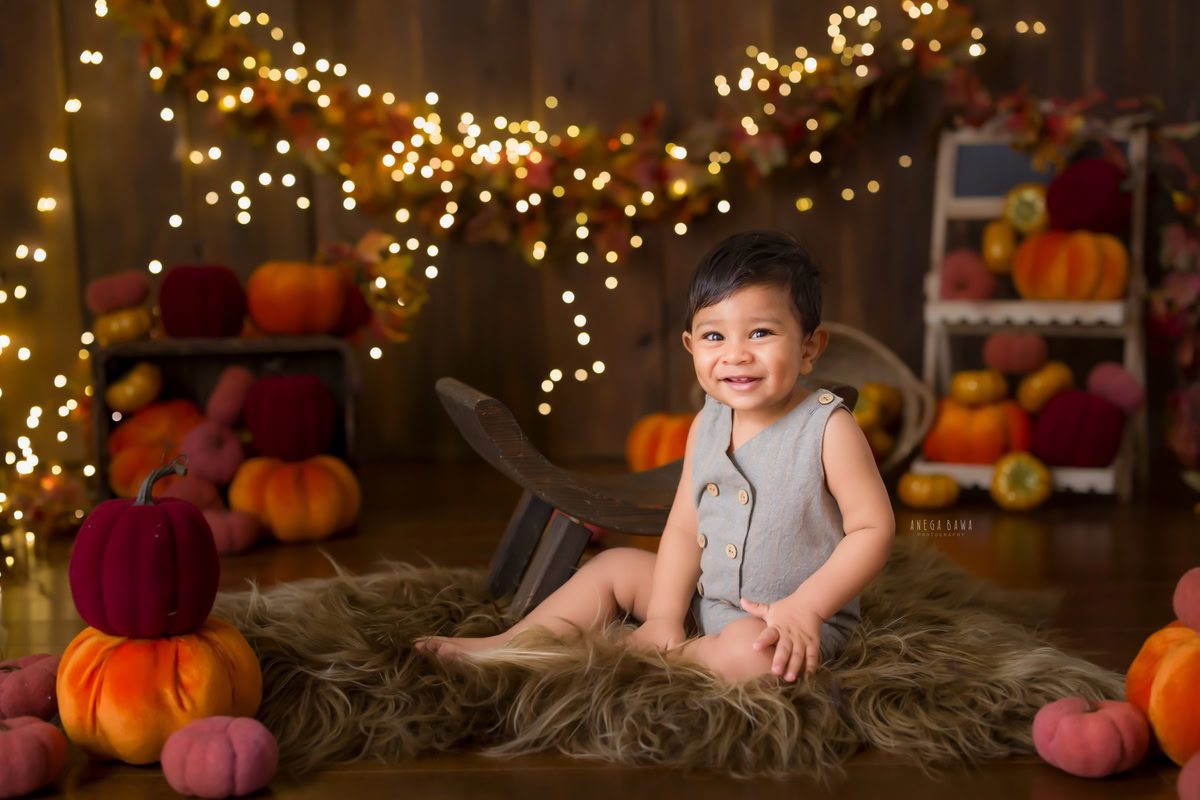 Captured against a warm brown backdrop, the 12-month-old boy sits comfortably on a plush brown rug, surrounded by the cozy glow of fairy lights. His playful spirit is evident as he interacts with an orange rust veggie soft toy, creating a charming scene during his baby pre-birthday photoshoot. Anega Bawa skillfully preserves this delightful moment, showcasing the joy of infancy in Delhi, Gurgaon, and Noida.
