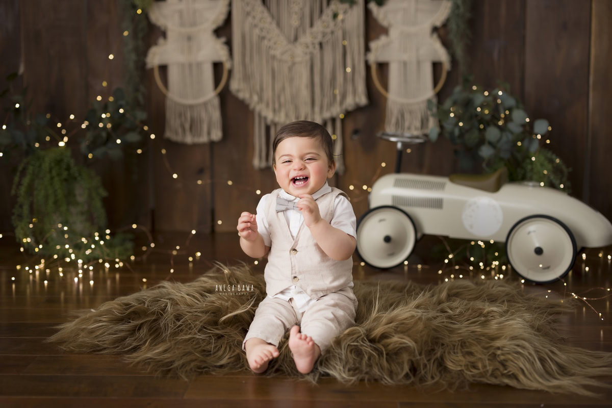 Captured beautifully in this toddler photoshoot by Anega Bawa in Delhi, Gurgaon, and Noida, the 10-month-old boy sits confidently on a brown rug, holding onto a toy scooter. The backdrop features a warm brown hue, complemented by green leafy pots and fairy lights, adding a touch of whimsy to the scene. With his playful demeanor and curious gaze, the boy exudes joy and innocence, making for a delightful photograph.