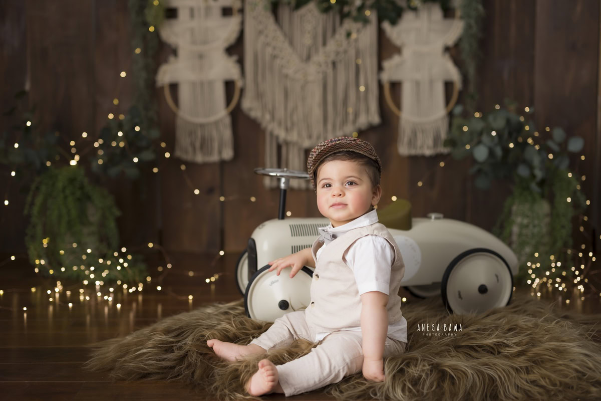 Delight in the precious moments as your little one, around 10 months to 1 year old, embarks on a charming sitter photoshoot with Anega Bawa Photography in Delhi and Gurgaon. Perched on a cozy brown rug, he explores his surroundings with gleeful curiosity, accompanied by a whimsical toy scooter. The soft glow of fairy lights adds a touch of magic to the scene, while a cute cap adds an extra dash of adorable charm. Capture these heartwarming memories as he sits amidst the beauty of childhood innocence and wonder.