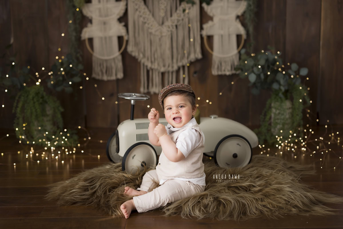 Embrace the joy of your little one's journey through childhood with Anega Bawa Photography in Delhi and Gurgaon. In this endearing sitter photoshoot, your child, aged around 10 months to 1 year, sits on a soft brown rug, exploring the world around him atop a charming toy scooter. Against a warm brown backdrop adorned with twinkling fairy lights and vibrant green pots, each moment is a treasure to be cherished. Let us capture these delightful memories as your child discovers the beauty of the world around him.