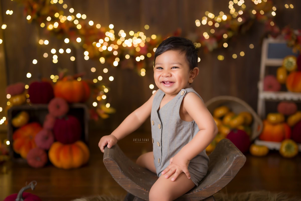 In the glow of twinkling fairy lights, the 10-month-old boy sits gracefully on a curvy chair against a warm brown backdrop. Beside him rests an adorable orange-brown veggie soft toy, adding a touch of whimsy to the scene. This captivating moment, captured during his baby pre-birthday photoshoot by Anega Bawa in Delhi, Gurgaon, and Noida, exudes charm and joy, celebrating the beauty of early childhood.