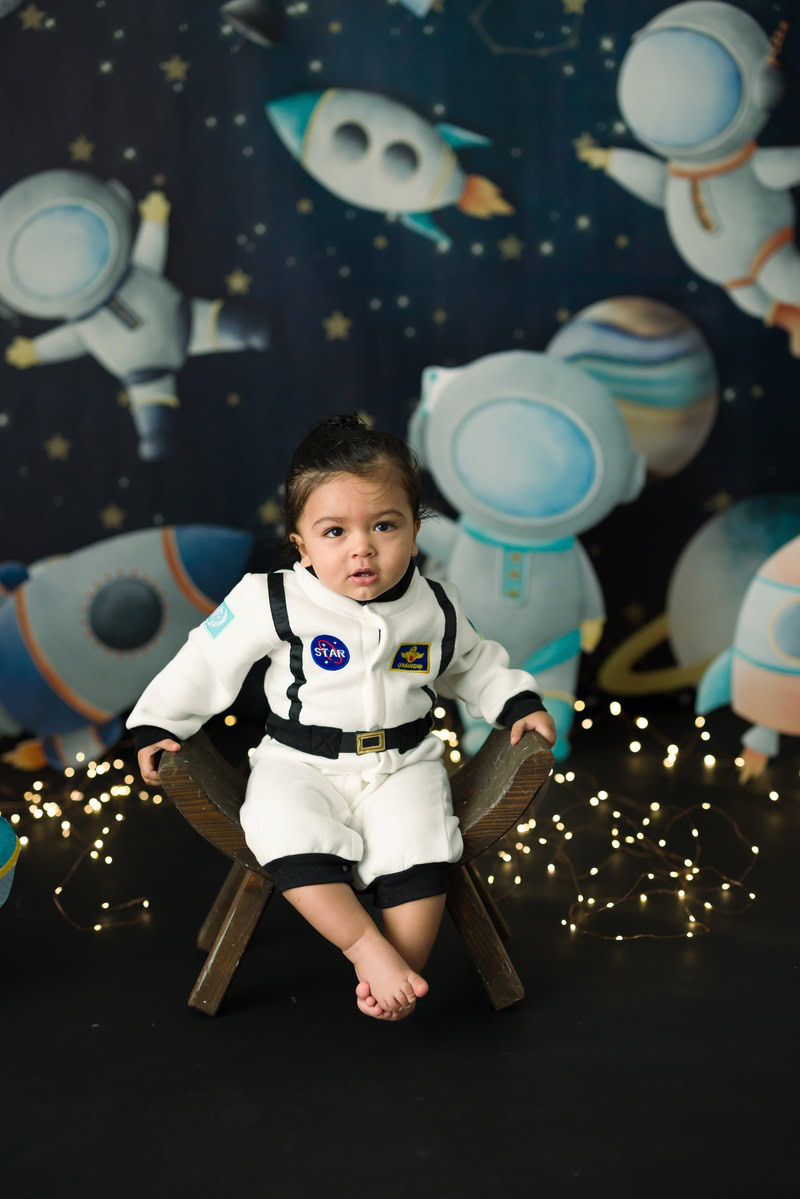 Delight in enchanting sitter photography by Anega Bawa in Delhi & Gurgaon. Your 10-month to 1-year-old sits on a curvy chair amidst a space backdrop with fairy lights.