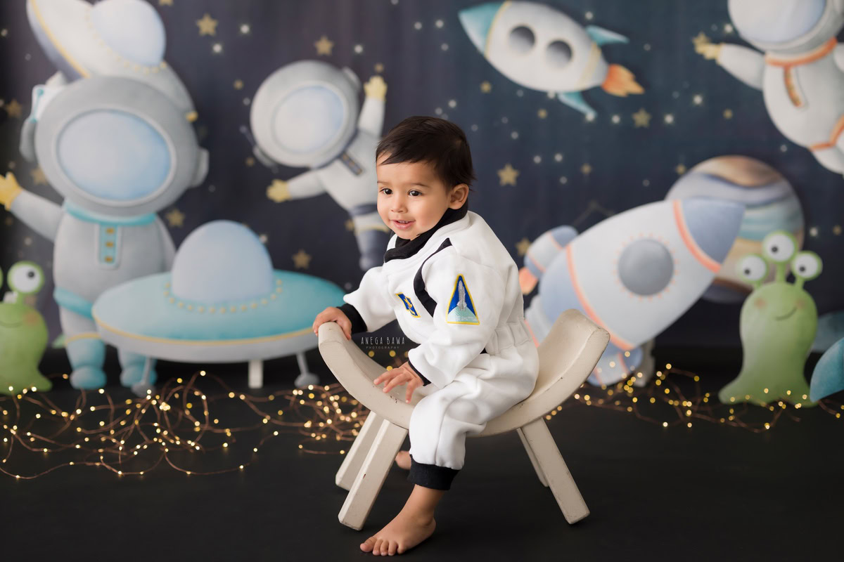 Boy sitting on a stylish curvy chair against a mesmerizing space backdrop, surrounded by twinkling fairy lights, capturing the magic of his journey from about 10 months to 1 year old during his first birthday photoshoot in Delhi by Anega Bawa, Gurgaon, Noida.